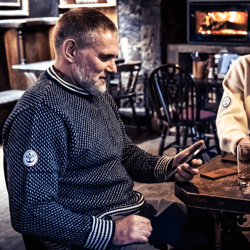 Man sitting in pub looking at mobile phone wearing Gunnerside Sweater 